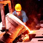 A factory worker wearing a helmet pours molten metal into molds, showcasing industrial metal casting.